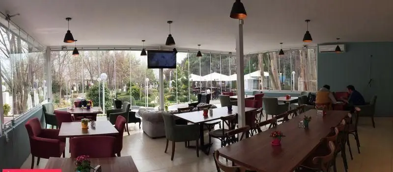 نمایی از فضای داخلی کافه و رستوران باشگاه مینی گلف با دکوراسیون شیک، میزهای چوبی و صندلی‌های راحت و پنجره‌های سراسری رو به فضای سبز و درختان.