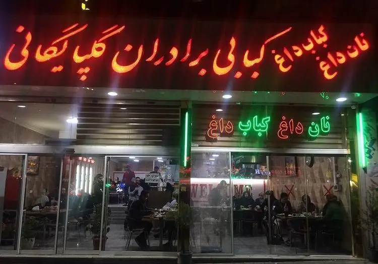 نمای بیرونی رستوران کبابی برادران گلپایگانی در شب، با تابلوی نئون کباب داغ، نان داغ و مشتریان در حال سرو غذا.