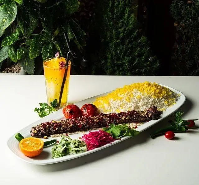 تصویری جذاب از یک پرس کباب خاص با برنج زعفرانی، گوجه کبابی و مخلفات تازه، همراه نوشیدنی خنک در کافه رستوران گرپ.