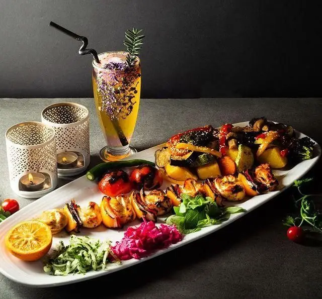 میز غذای شیک در کافه رستوران گرپ: شامل سیخ‌های کباب، سبزیجات گریل، سالاد، ترشی و نوشیدنی خاص با شمع‌های دکوراتیو.