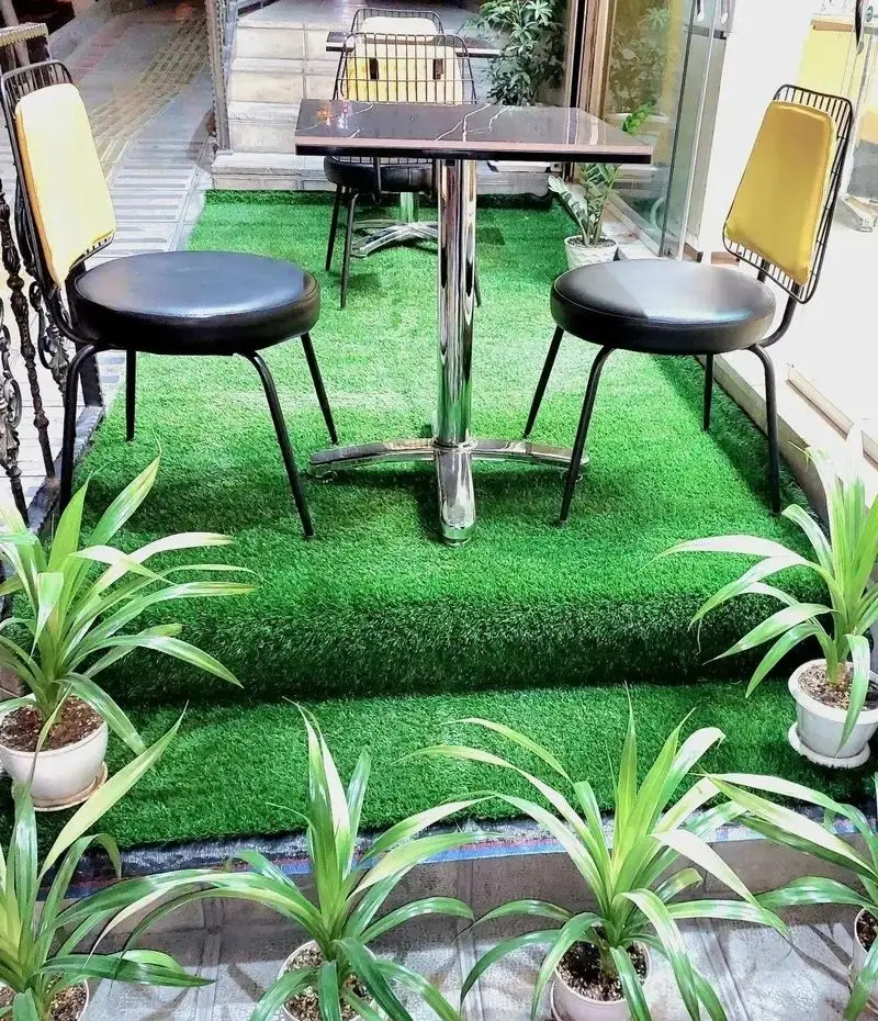 فضای بیرونی جذاب پیتزا علامت سؤال با چمن مصنوعی، میز طرح سنگ و صندلی‌های مدرن سیاه و زرد، آماده پذیرایی از مشتریان.