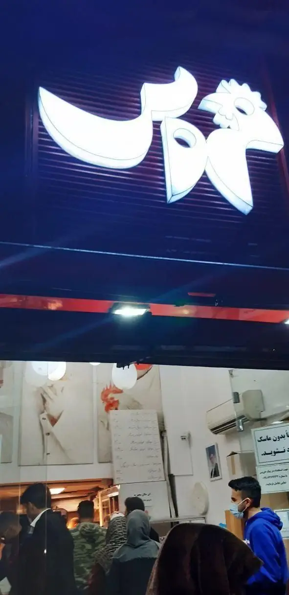 نمای شب از تابلوی نئون فست فود خروس و فضای داخلی رستوران با صف مشتریان که ماسک زده‌اند و تابلوی "ورود بدون ماسک ممنوع".