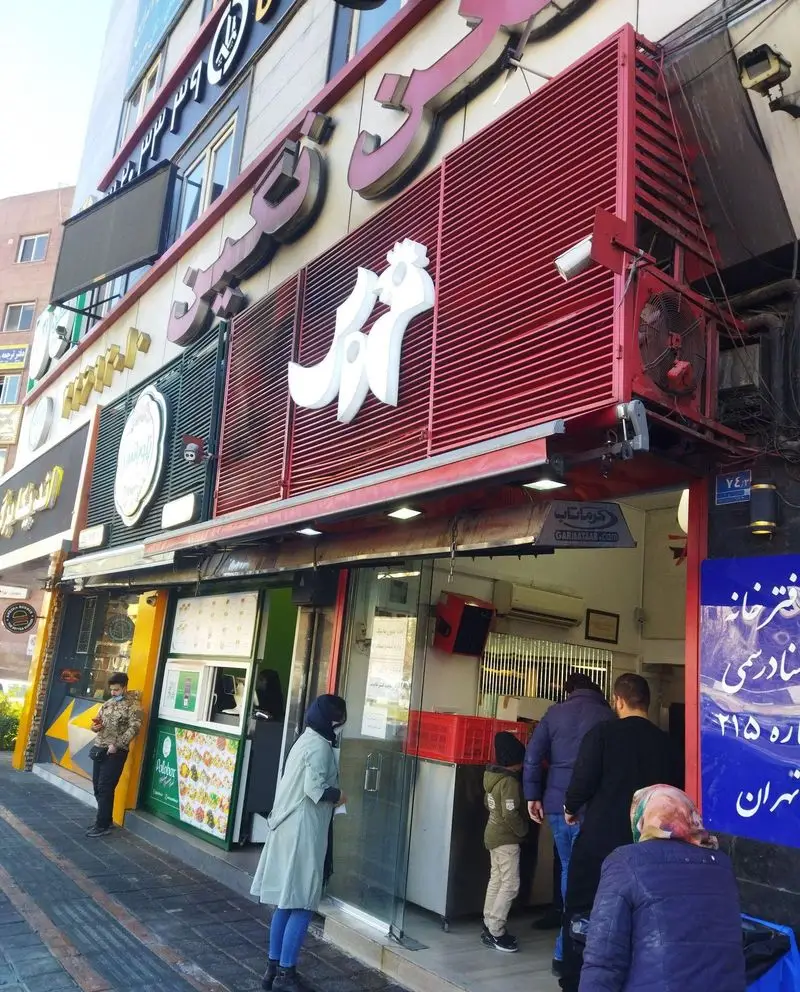 نمایی جذاب از سردر فست فود خروس در تهران با لوگوی سفید رنگ و مشتریان منتظر سفارش غذای آماده.