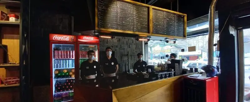 رستوران میخوش: فضای داخلی شیک با کارکنان ماسک‌زده پشت کانتر، منوی غذا و نوشیدنی روی تخته سیاه، و یخچال‌های پر از نوشیدنی.