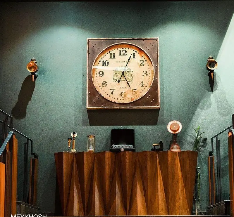 دکوراسیون داخلی شیک و خاص رستوران میخوش با دیوار سبز آبی، ساعت دیواری بزرگ وینتیج و کنسول چوبی مدرن و اکسسوری‌های کلاسیک.