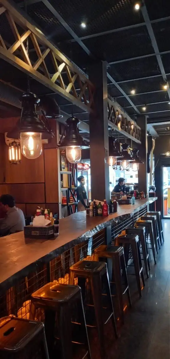 تصویر دکوراسیون داخلی جذاب رستوران میخوش با میز بار بلند، صندلی‌های فلزی، نورپردازی صنعتی و سقف چوبی، محیطی مدرن و دنج.