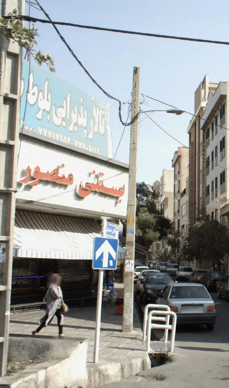 نمای بیرونی مغازه بستنی منصور و تالار پذیرایی بلوط در خیابانی شلوغ از یک شهر ایرانی، با خودروها و رهگذران در روز روشن.