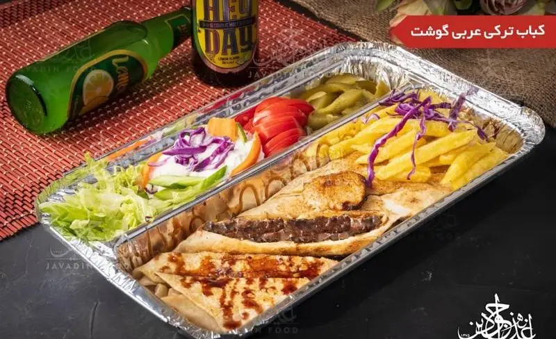 پک ساندویچ کباب ترکی گوشت اغذیه جوادین همراه با سیب‌زمینی سرخ کرده، خیارشور، سالاد و گوجه، نوشیدنی در پس زمینه.