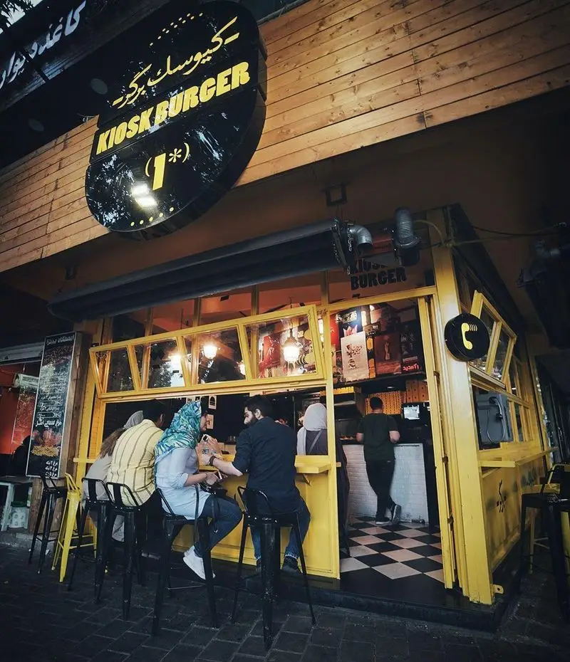 کیوسک برگر: نمای بیرونی فست فود با دکوراسیون زرد جذاب و مشتریان در حال لذت بردن از همبرگرهای خوشمزه.