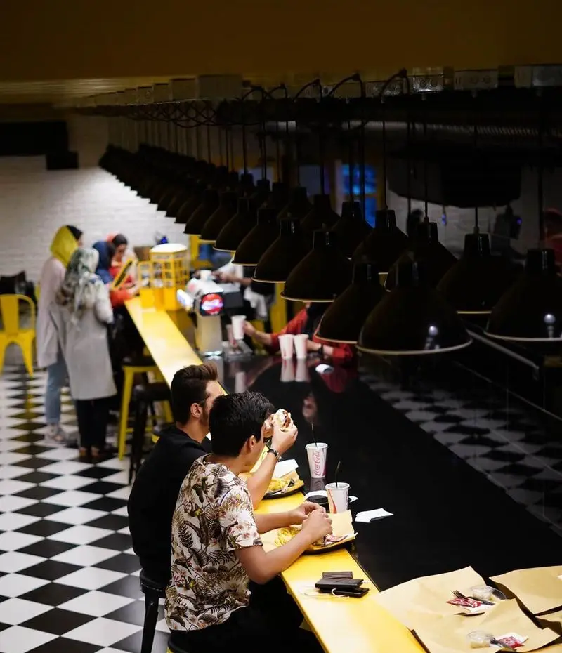 مشتریان در حال لذت بردن از غذاهای خوشمزه و برگرهای کیوسک برگر در فضایی مدرن و دلنشین با طراحی منحصر به فرد و نورپردازی خاص.