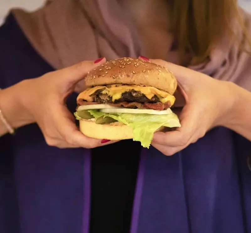 تصویری نزدیک از دستان یک نفر که یک چیزبرگر بزرگ و آبدار با پنیر ذوب شده، بیکن و سبزیجات تازه از کیوسک برگر را نگه داشته است. طعم بی‌نظیر.