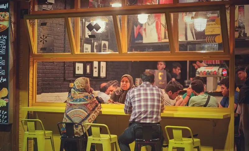 فست فود کیوسک برگر، مشتریان در کنار پیشخوان زرد رنگ در شب. منوی متنوع غذاها شامل پیتزا و مرغ در محیطی دوستانه