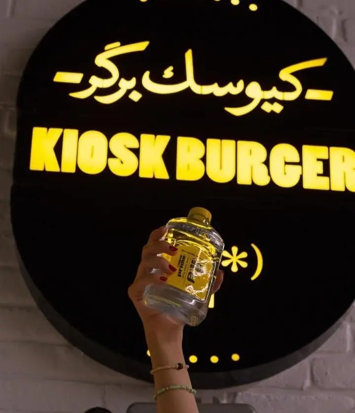 تصویر تابلو جذاب برند فست فود کیوسک برگر با فونت زرد روشن روی زمینه مشکی، همراه با دست فردی که بطری نوشیدنی نگه داشته.