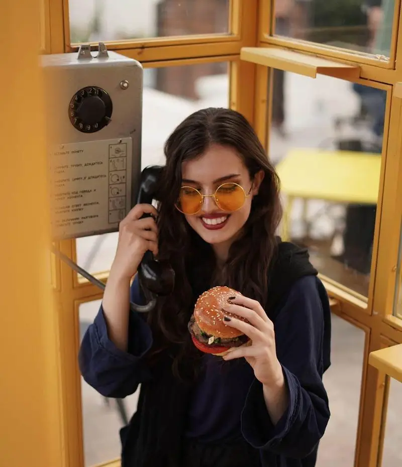 زنی خندان با عینک زرد در باجه تلفن در حال لذت بردن از یک برگر خوشمزه کیوسک برگر، تجربه‌ای عالی و متفاوت برای غذای شما.
