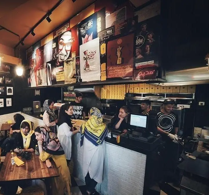 نمایی از داخل رستوران فست فود کیوسک برگر با مشتریان در حال ثبت سفارش و پرسنل مشغول خدمت رسانی. دکوراسیون هنری و منحصر به فرد.