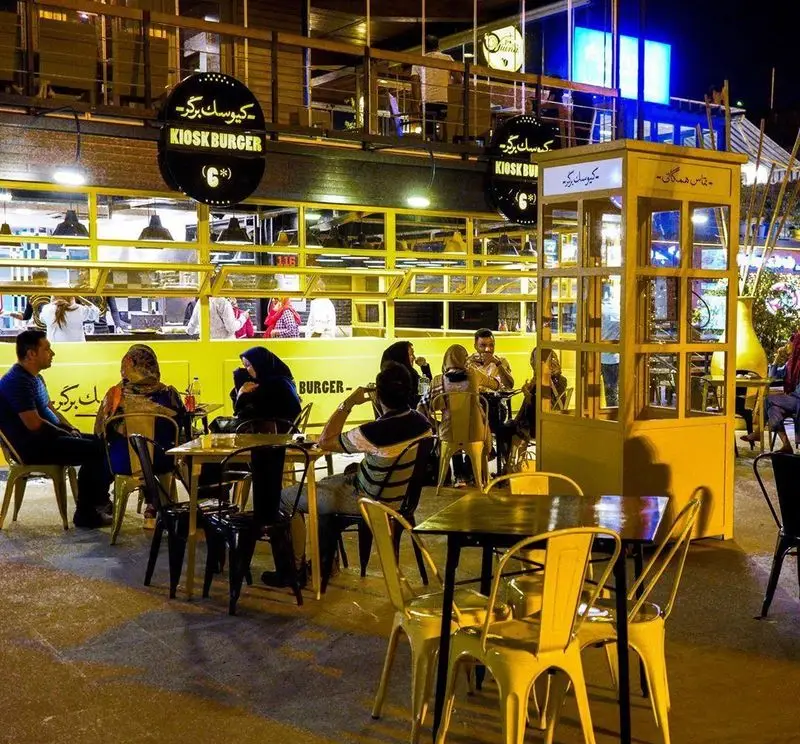 کیوسک برگر: نمایی از رستوران فست فود با طراحی مدرن و فضای باز شلوغ در شب، مشتریان در حال صرف غذا و نوشیدنی.