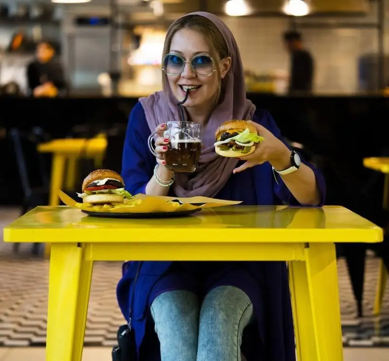 زنی جوان و خندان با عینک و حجاب، در حال نوشیدن و خوردن همبرگر خوشمزه در محیط دلنشین کیوسک برگر، تجربه‌ای لذت‌بخش از فست فود