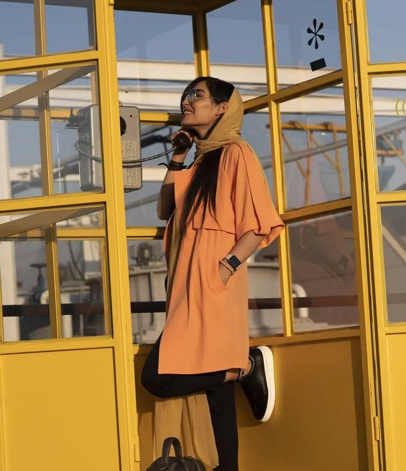 تصویر زنی شاد با لباس نارنجی و شال زرد، در حال استفاده از تلفن عمومی در کیوسک زردرنگ، متناسب با حال و هوای کیوسک برگر.