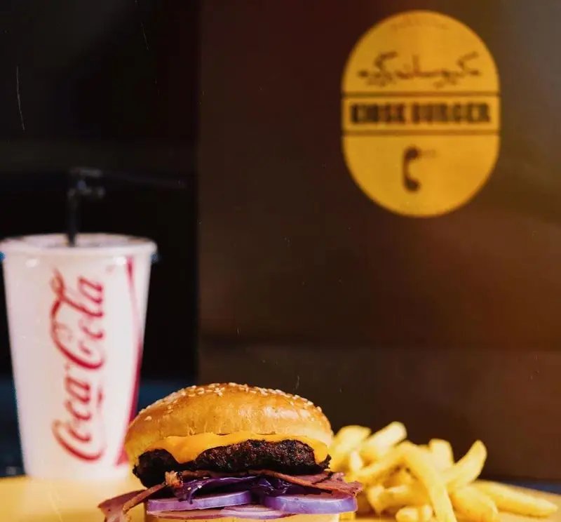 تصویری از یک همبرگر خوشمزه با پنیر، پیاز و مخلفات، در کنار سیب زمینی سرخ کرده و نوشابه کوکاکولا در محیط رستوران کیوسک برگر.