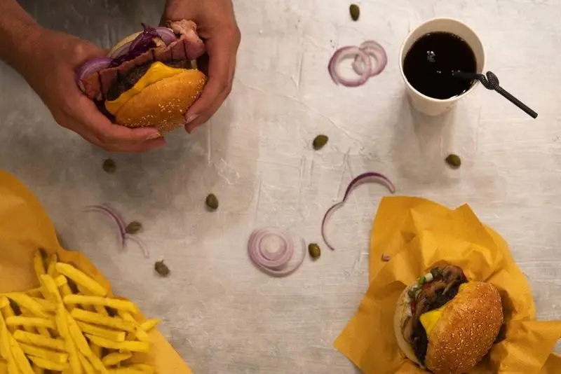 سفارش دو برگر، سیب‌زمینی و نوشیدنی سرد از کیوسک برگر. برگرها شامل پنیر، بیکن و پیاز قرمز. تجربه یک وعده غذایی لذیذ فست فود.