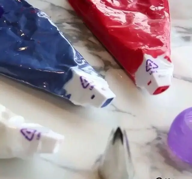 لوازم قنادی دست ساز: قیف‌های خامه پزی آبی، قرمز و سفید پرشده از رنگ خوراکی، در کنار ماسوره استیل؛ ابزاری ایده‌آل برای تزئین کیک و شیرینی.