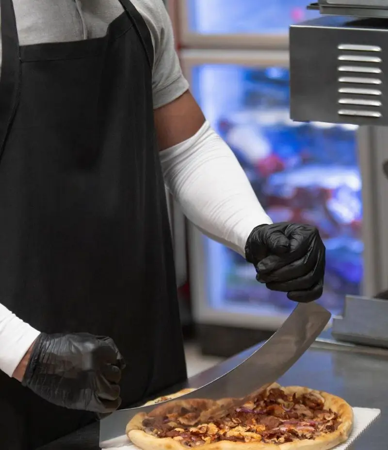 آشپز فست فود شیلا در حال برش دقیق پیتزای خوشمزه و داغ با روکش گوشت در محیط بهداشتی رستوران برای ارائه بهترین طعم.