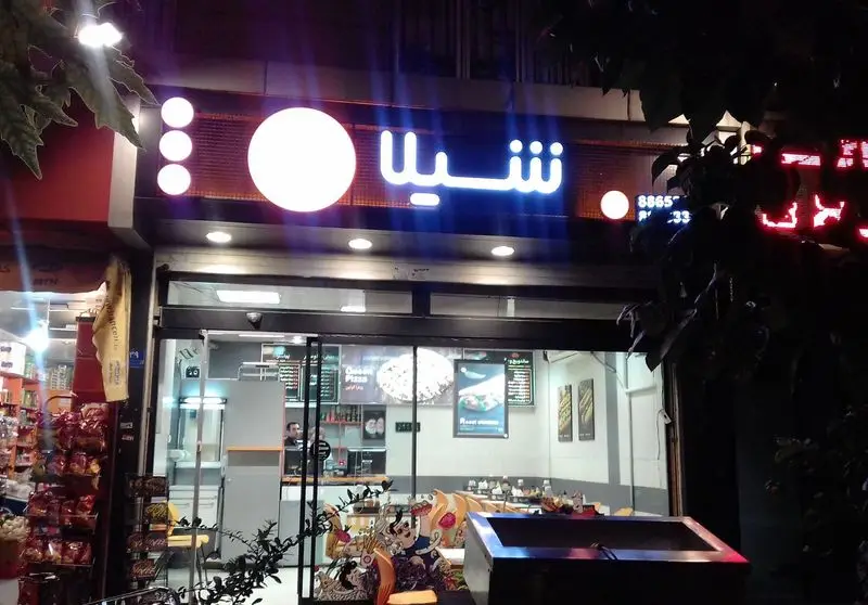 نمای خارجی روشن فست فود شیلا در شب با تابلوی نئون جذاب و واضح. این رستوران با فضای داخلی مدرن و ویترین شیشه ای برای مشتریان آماده است.