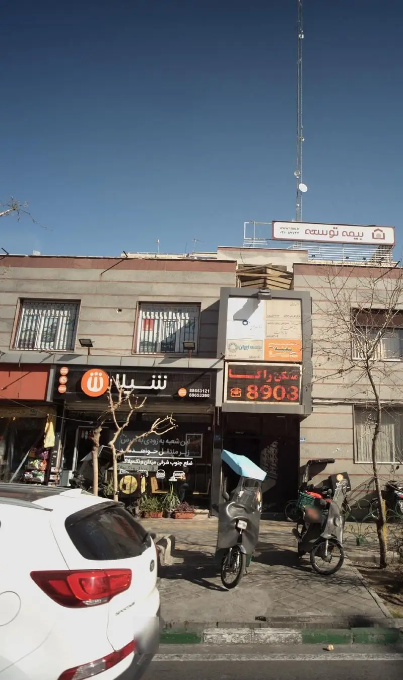 تصویر نمای بیرونی شعبه فست فود شیلا، با تابلوی مشخص و موتورهای پیک آماده ارسال غذا، در یک روز روشن و شلوغ در شهر.