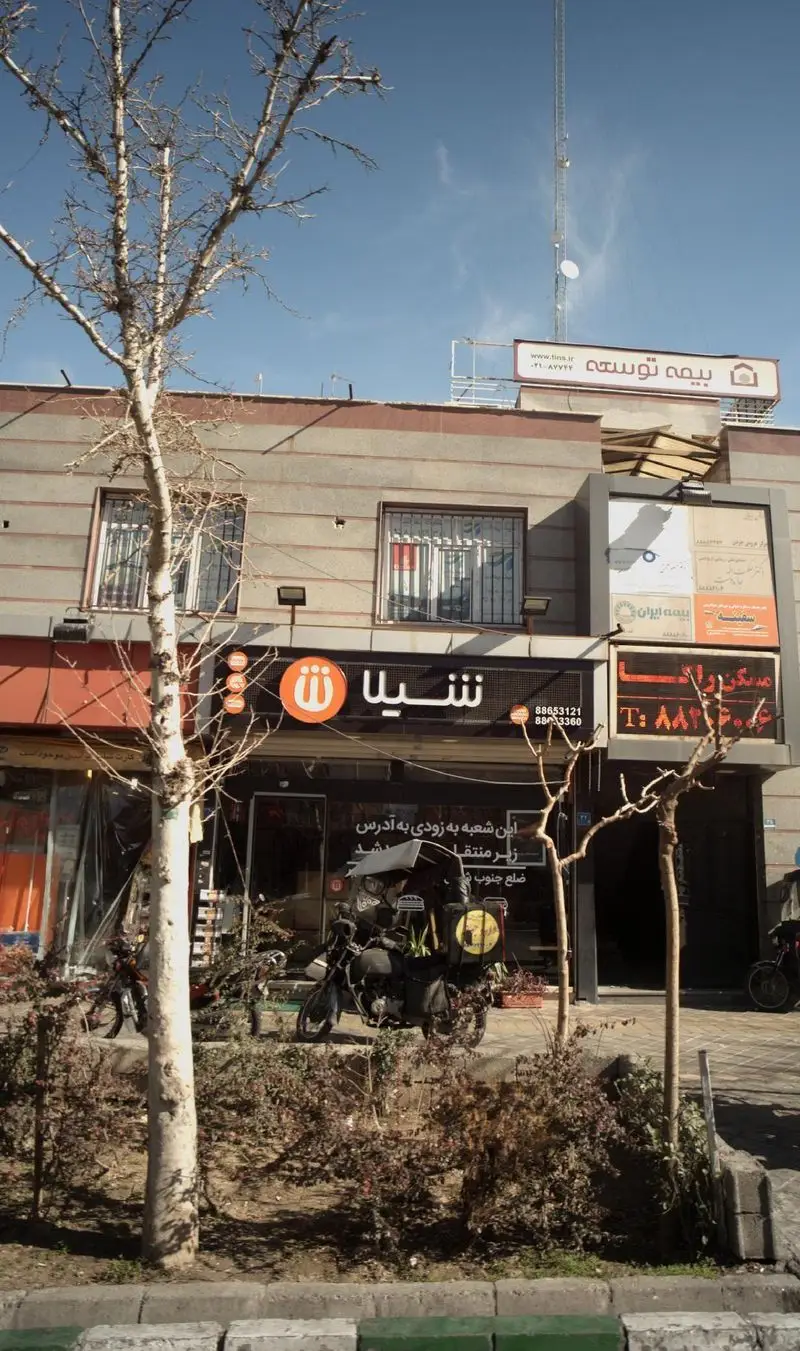 نمایی از شعبه فست فود شیلا در تهران، شامل نمای بیرونی رستوران، لوگو و موتورسیکلت تحویل غذا. سفارش آنلاین و حضوری.