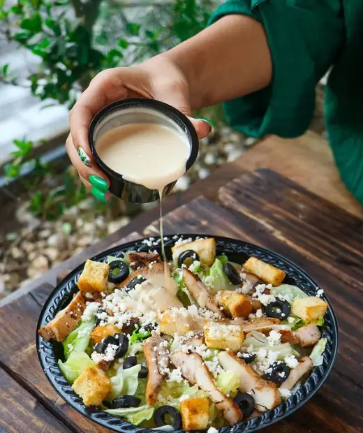 ریختن سس مخصوص روی سالاد سزار مرغ شیلا شامل کاهو، تکه‌های مرغ گریل، زیتون سیاه، کروتون و پنیر فتا. غذای سالم و لذیذ.