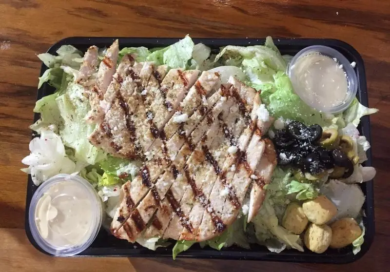 نمای نزدیک از سالاد مرغ گریل لذیذ رستوران پروفسور: سینه مرغ کبابی، کاهو، زیتون سیاه و سبز، کروتون و سس در ظرف بیرون‌بر.