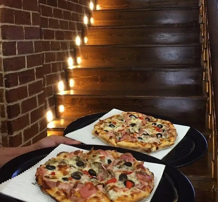 پیتزاهای خوشمزه رستوران پروفسور، شامل پیتزای ژامبون، قارچ و زیتون، آماده سرو در محیط دلنشین با پله‌های چوبی و دیوار آجری.