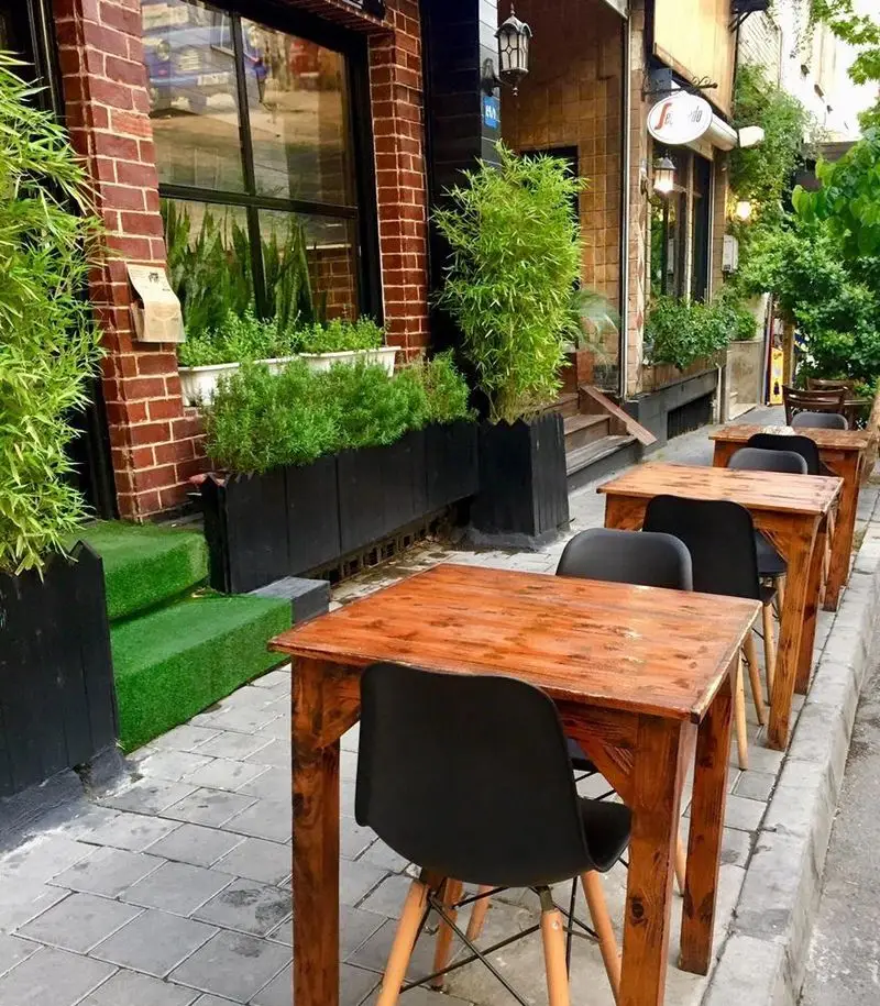 نمای بیرونی رستوران پروفسور با میز و صندلی‌های چوبی در فضای باز پیاده‌رو و گلدان‌های سرسبز، محیطی دلنشین برای صرف غذا.