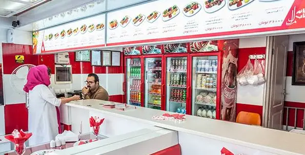 تصویری از فضای داخلی رستوران خانه کباب ملل، شامل پیشخوان سفارشات، مشتری، کارمند، منوی کباب‌ها و نوشیدنی‌های خنک در یخچال.