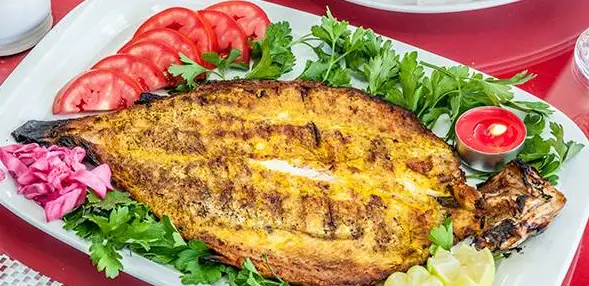 سفره‌ای رنگین از ماهی کباب شده زردرنگ طلایی، با برش‌های گوجه‌فرنگی تازه، سبزیجات معطر و ترشی برای تجربه‌ای دلنشین در خانه کباب ملل.