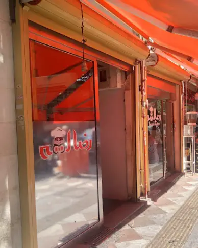 نمای بیرونی مغازه و ورودی فست فود رضا لقمه با سقف نارنجی، شیشه‌های براق و آرم آشپز، در یک روز آفتابی.