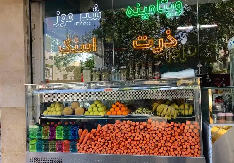 نمای کسب و کار آبمیوه و بستنی ملو، با تابلوهای نئون شیر موز، ویتامینه و ذرت، و پیشخوان پر از میوه های تازه و هویج.