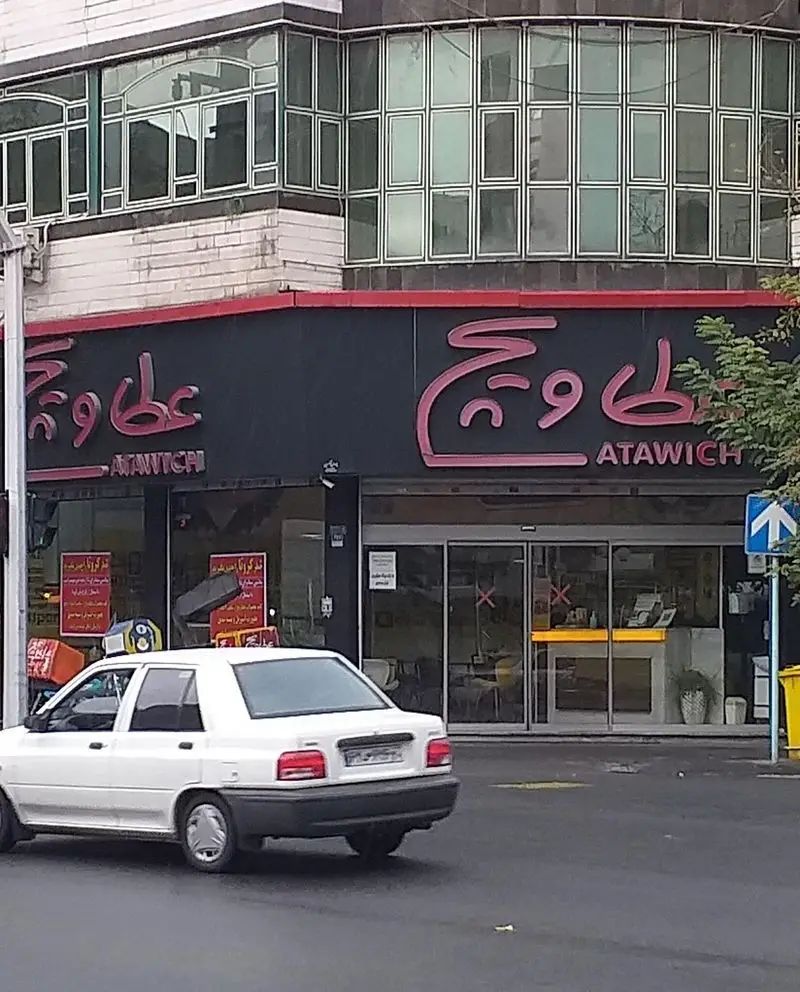 نمای خارجی شعبه عطاویچ: تابلوی صورتی رنگ برند بر پس زمینه مشکی، همراه با ورودی شیشه‌ای و یک خودروی سفید در حال عبور از خیابان.