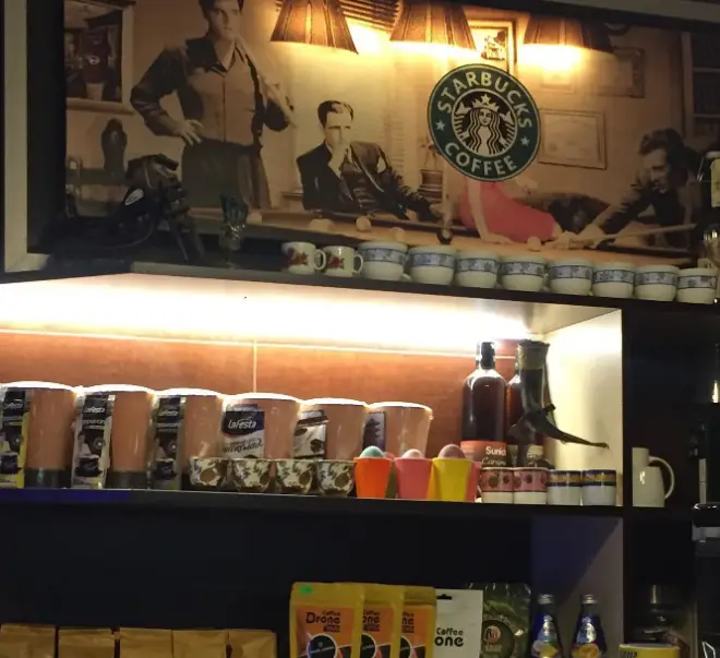 تصویری از قفسه محصولات کافه آسا: دکوراسیون جذاب با طرح‌های قدیمی، لوگوی استارباکس، قهوه‌های بسته‌بندی شده LaFesta و Drone و نوشیدنی‌ها.