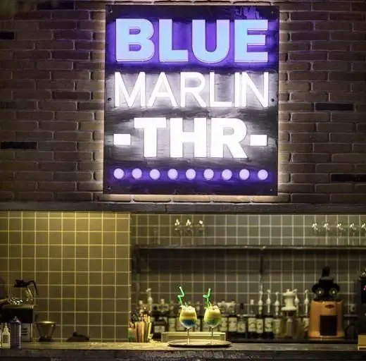 کافی شاپ بلومارلین: تابلوی نئونی "BLUE MARLIN THR" روی دیوار آجری، بار مجهز با نوشیدنی‌های متنوع و فضای دلنشین.
