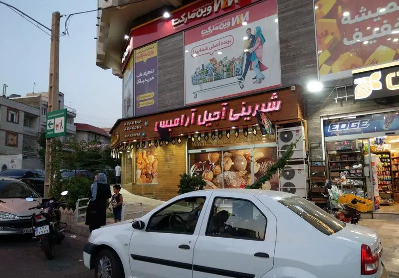 نمای بیرونی فروشگاه شیرینی و آجیل آرامیس در یک خیابان شلوغ شهری، با تابلوهای نئون درخشان و ویترین پر از شیرینی و نان تازه.