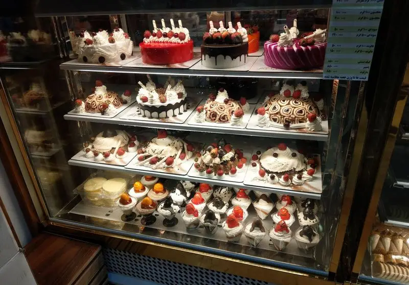 نمایی از ویترین لوکس شیرینی و کیک های مجلسی و تازه قنادی آرامیس، شامل انواع کیک تولد، دسر، کاپ کیک با تزیینات میوه و خامه.
