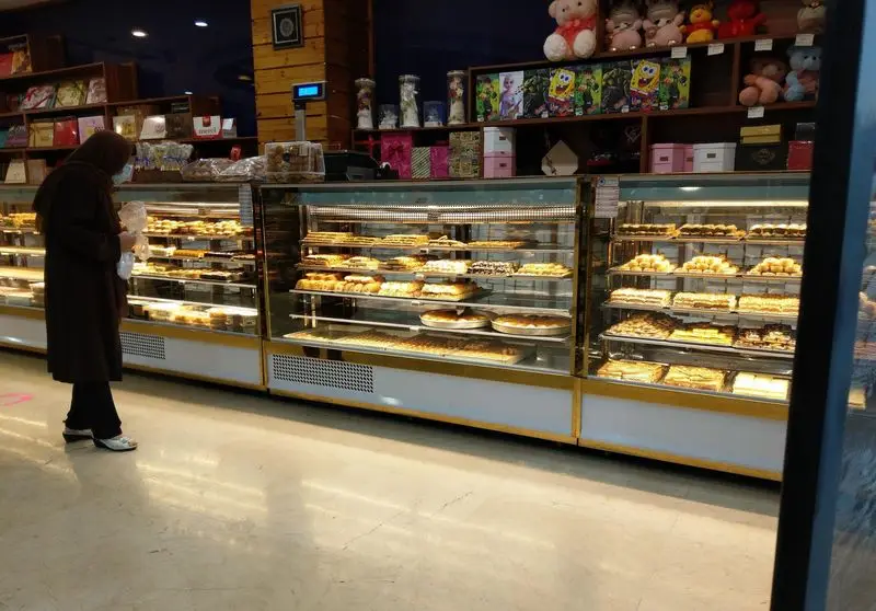 تصویری از مشتری در قنادی و آجیل آرامیس، در حال مشاهده ویترین‌های بزرگ پر از انواع شیرینی‌های تازه و خوشمزه سنتی و مدرن برای خرید.