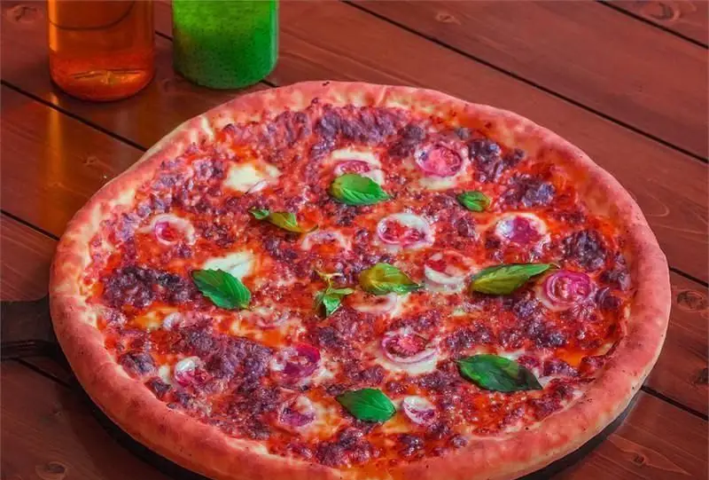 پیتزای جذاب و خوشمزه از پیتزایی بلا، با خمیر طلایی، سس قرمز، پنیر، پیاز و ریحان تازه روی میز چوبی، آماده سفارش.