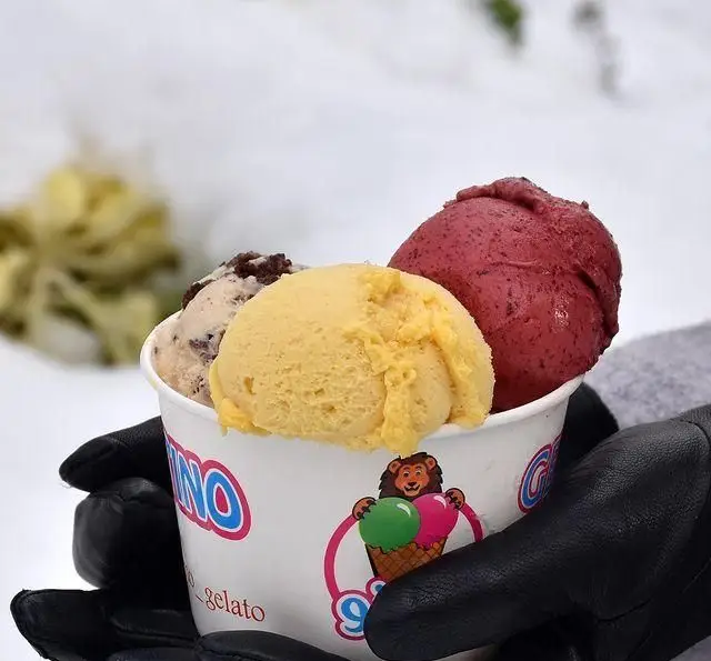 سه اسکوپ جلاتوی رنگارنگ از کافه جلاتو متینو، شامل شکلاتی، انبه و توت، در دستکش مشکی در منظره برفی زمستان.