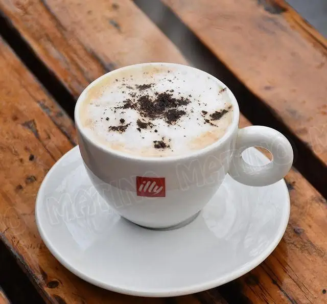 کاپوچینو داغ ایلی با پودر کاکائو در فنجان سفید و نعلبکی روی میز چوبی، نوشیدنی دلپذیر از کافه جلاتو متینو.
