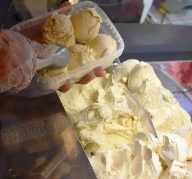 در کافه جلاتو متینو، دستان ماهر با دستکش مشغول اسکوپ زدن بستنی جلاتو باکیفیت و تازه به رنگ کرم/پسته در ظرف شفاف هستند.