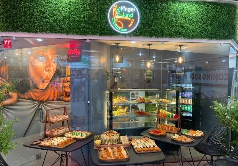 نمای جذاب کافه فینگر لند با لوگوی نئون سبز، ویترین غذاهای انگشتی متنوع، دسر و ساندویچ‌های کوچک آماده سرو.