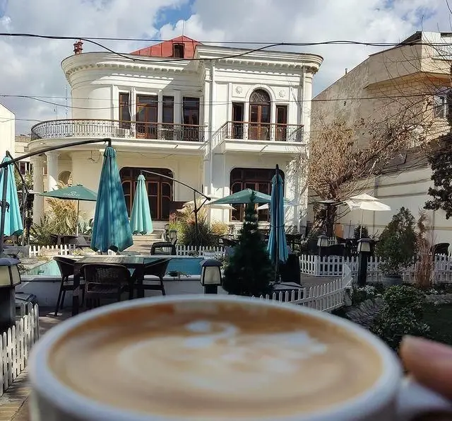 نمایی دلنشین از کافه مرکوری با معماری سفید و سقف قرمز، حیاطی با چترهای آبی، میز و صندلی‌ها و قهوه لذیذ با لاته آرت