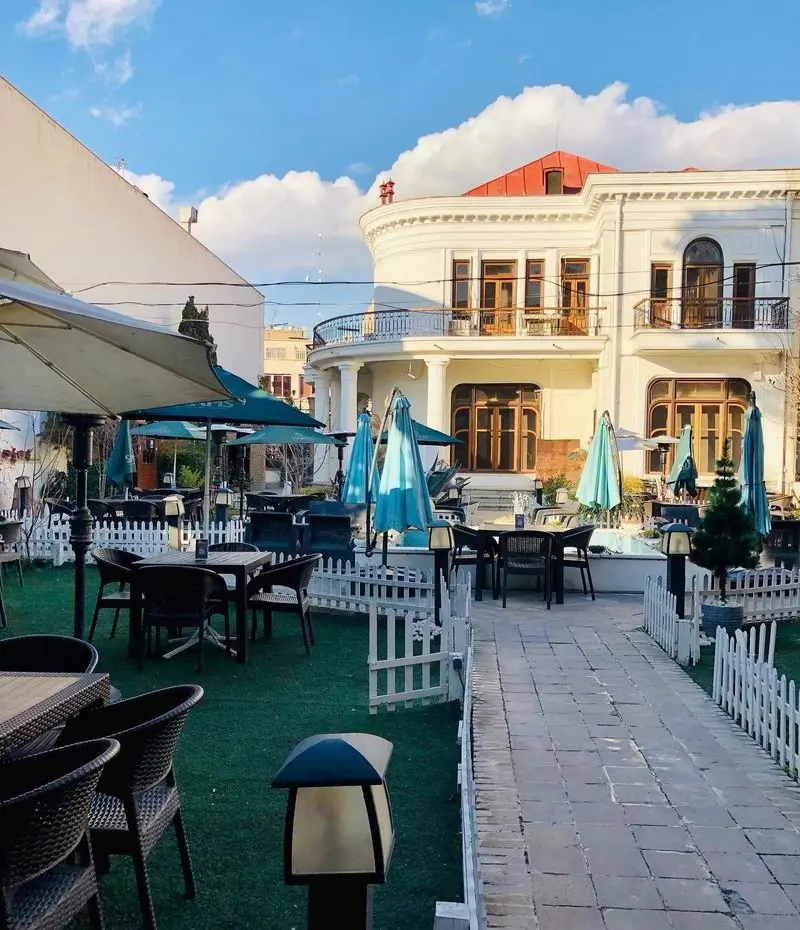 تصویری از فضای باز زیبا و دلنشین کافه مرکوری با میز و صندلی‌های شیک، چترهای آبی و سبز و مسیر سنگفرش شده و ساختمان سفیدرنگ